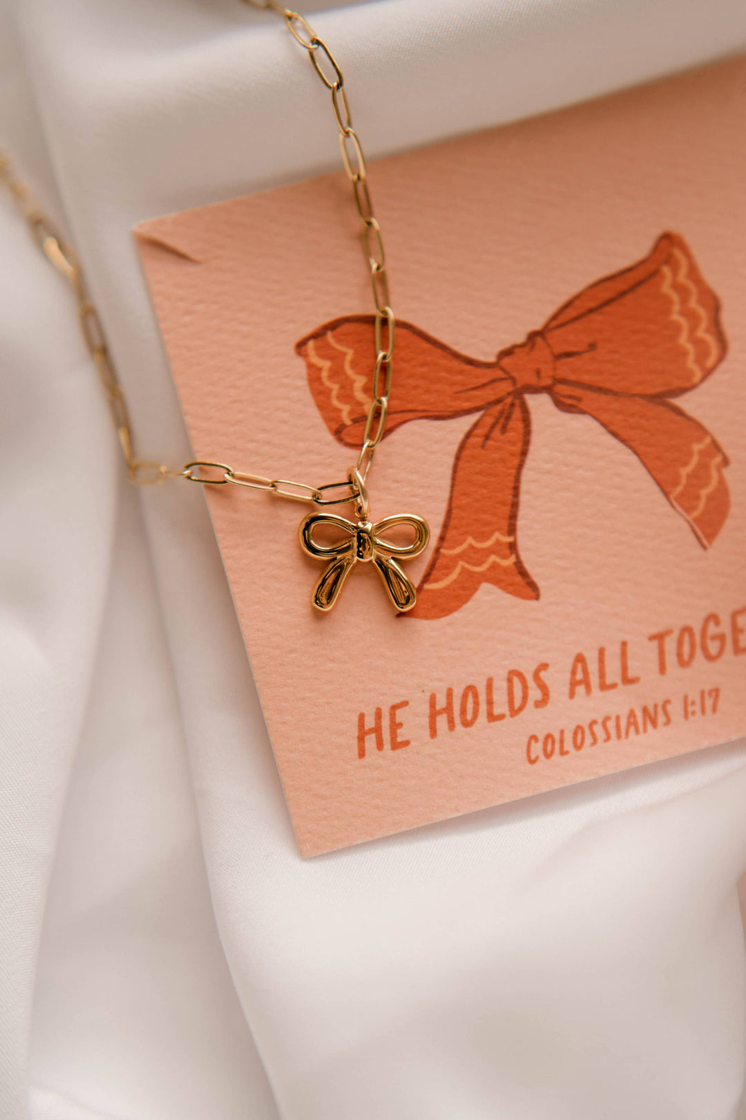 Gold necklace with a bow pendant on a pink card with text