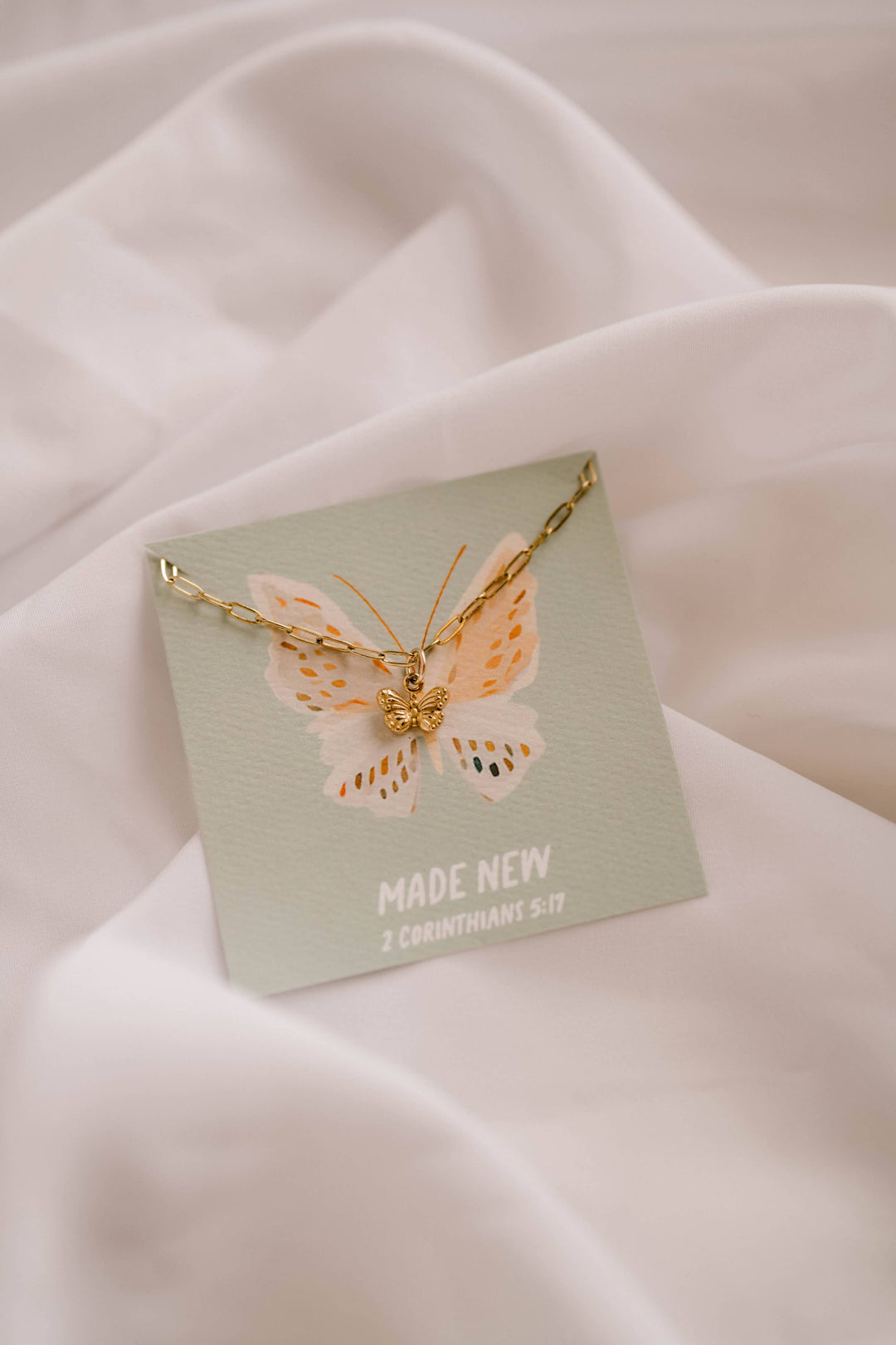 Gold butterfly necklace on a green card with 'MADE NEW' text, against a soft pink fabric background