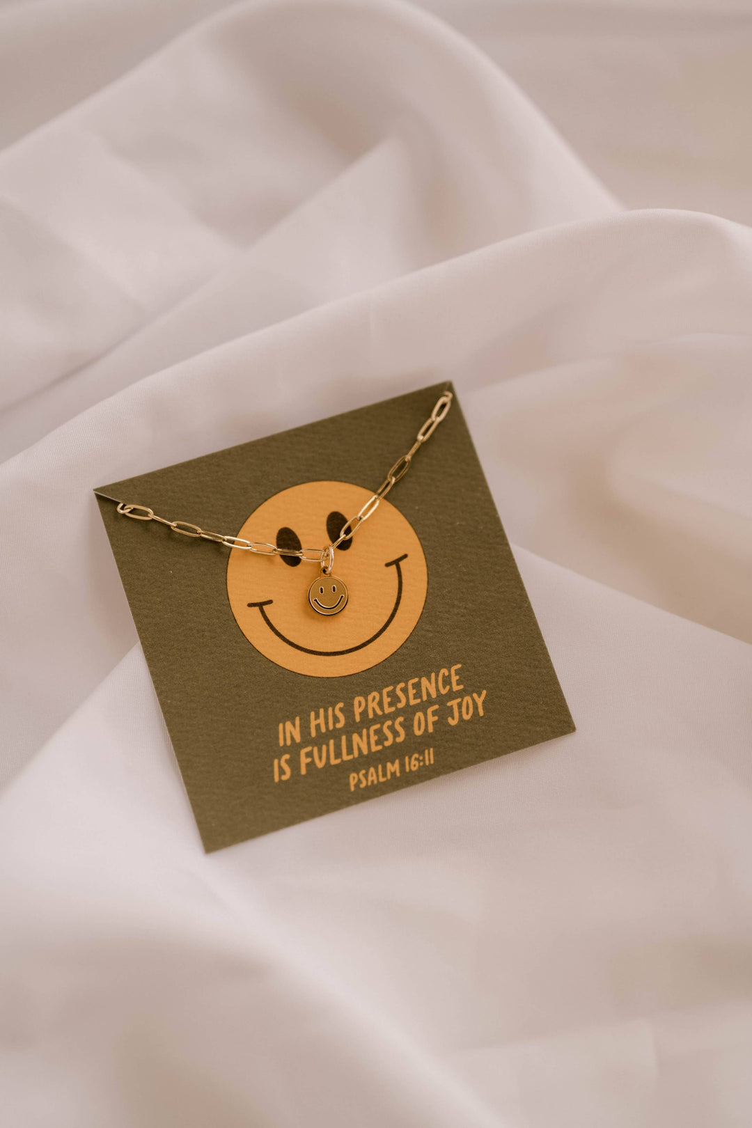 Gold necklace with smiley face pendant on a green card with text, placed on a white fabric background