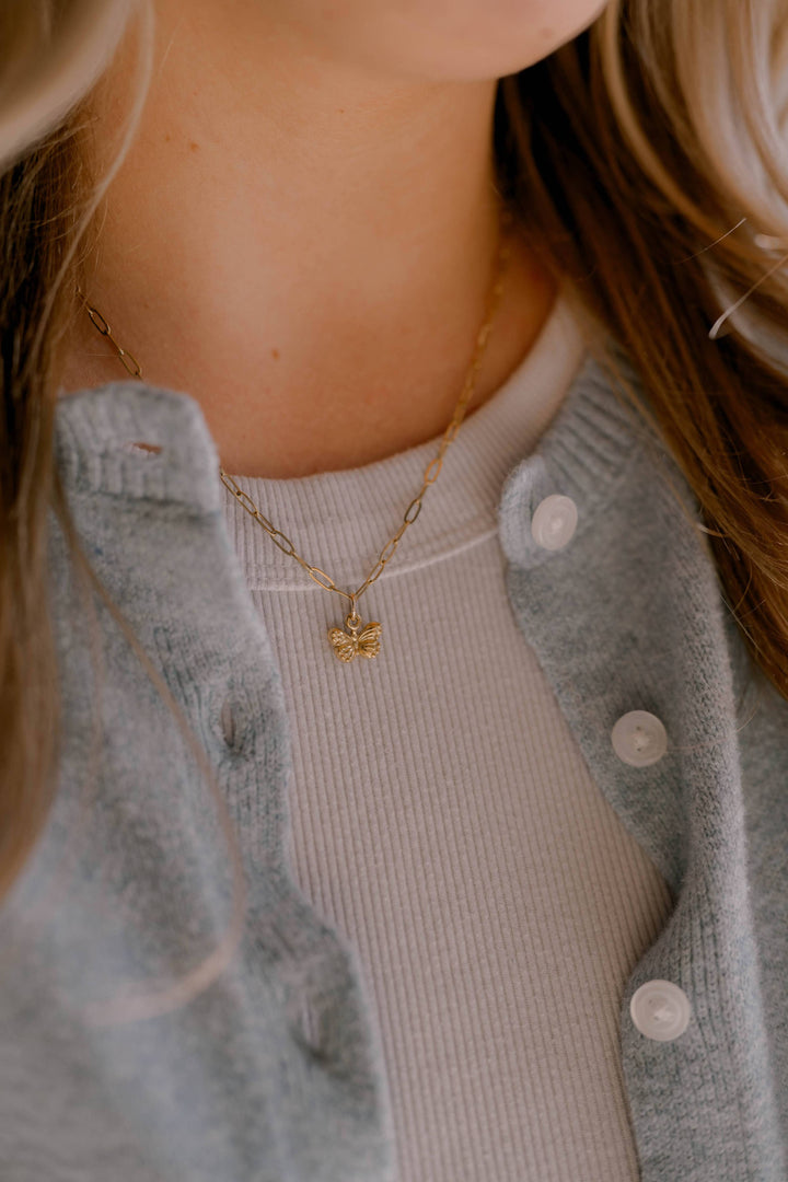 Person wearing a gold necklace with a heart pendant, wearing a gray cardigan over a beige top.