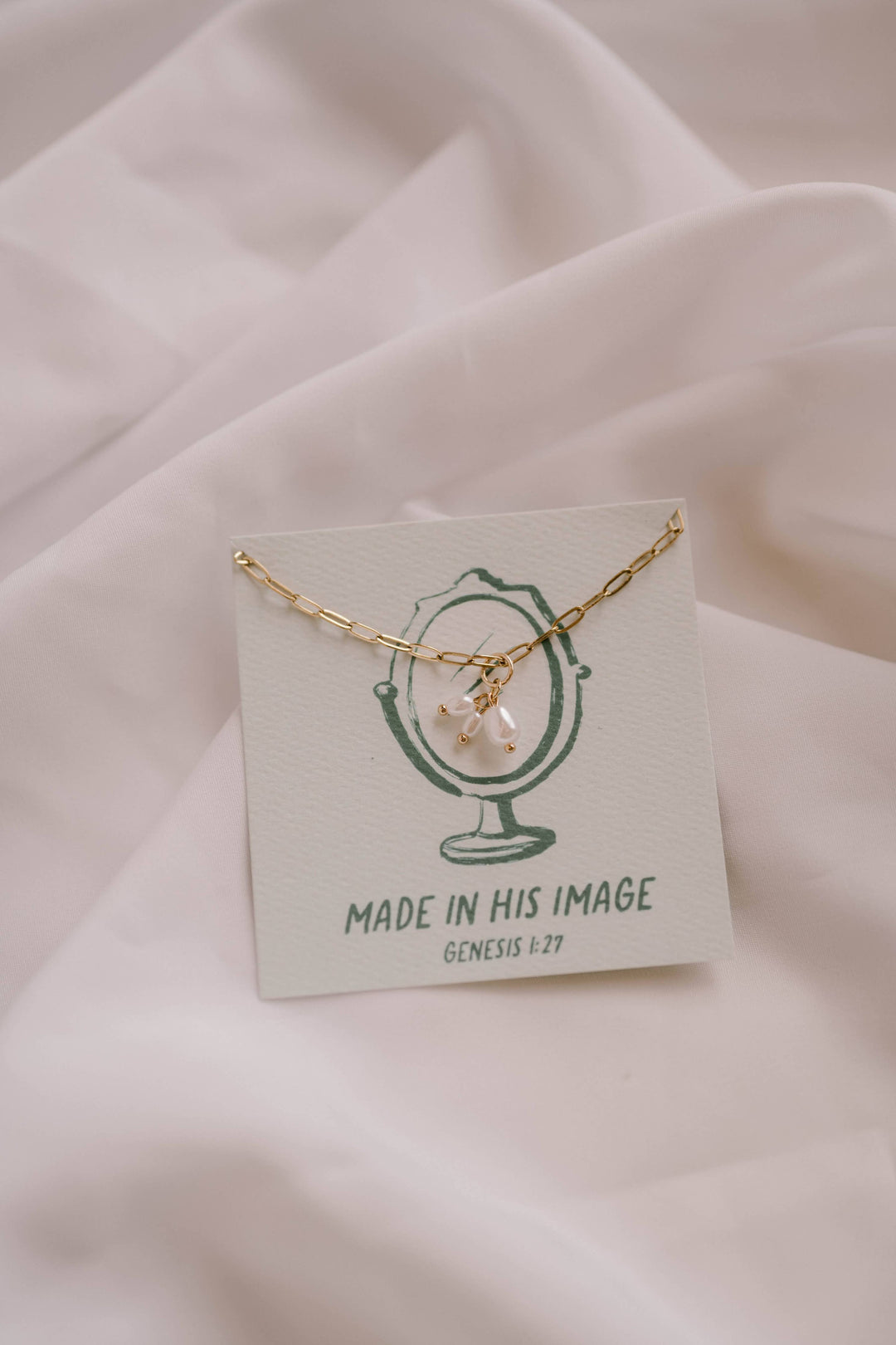 Gold necklace with a pink pendant on a card with text, placed on a soft pink fabric background.