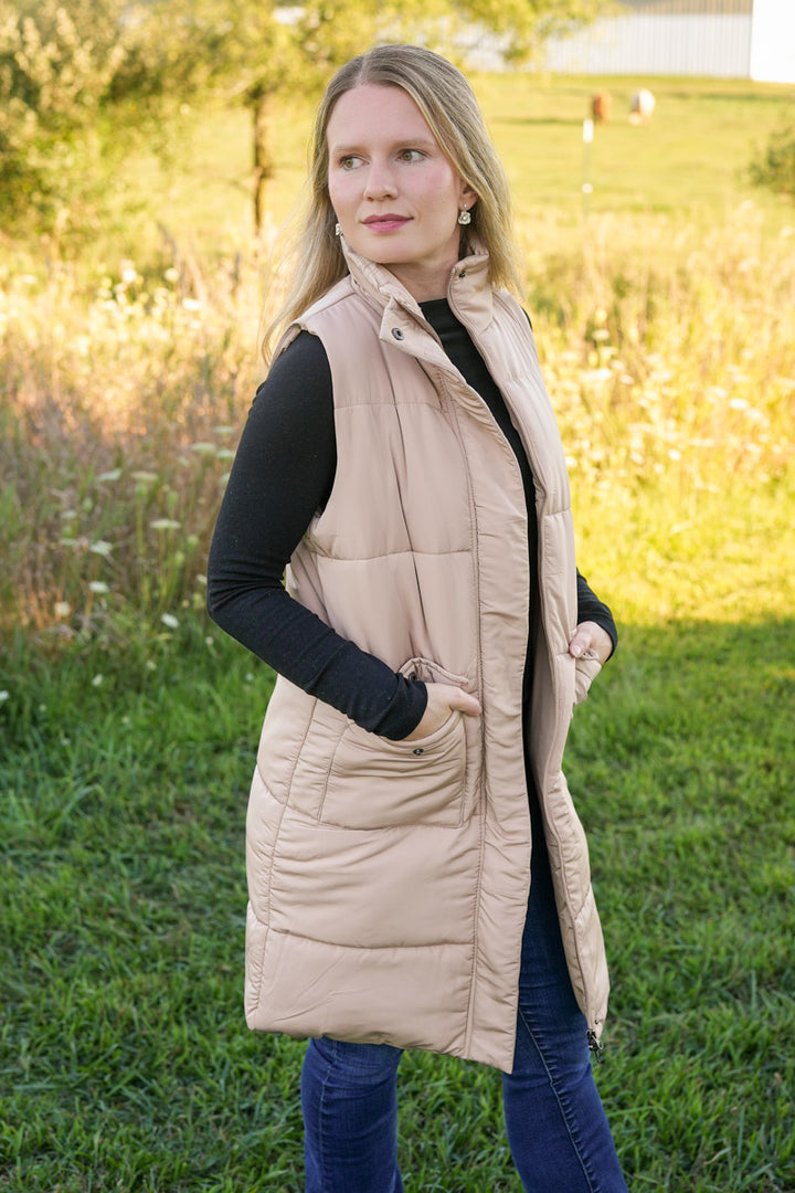 Woman wearing a long beige puffer vest with pockets in a grassy outdoor setting