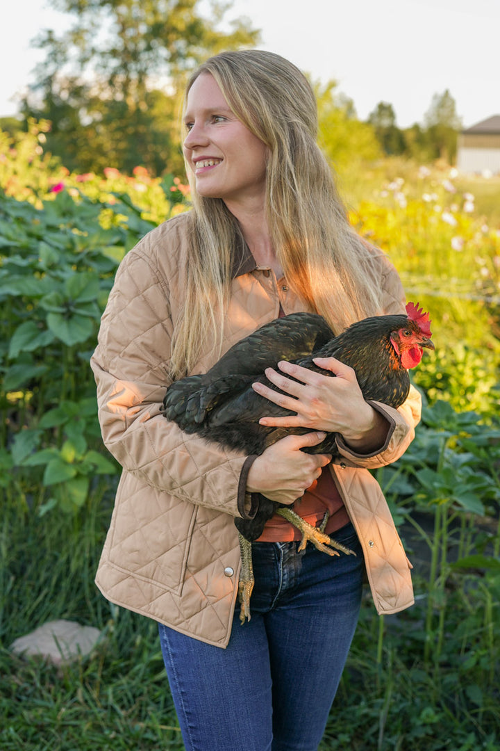 Woman wearing a tan quilted barn jacket with contrast collar holding a chicken