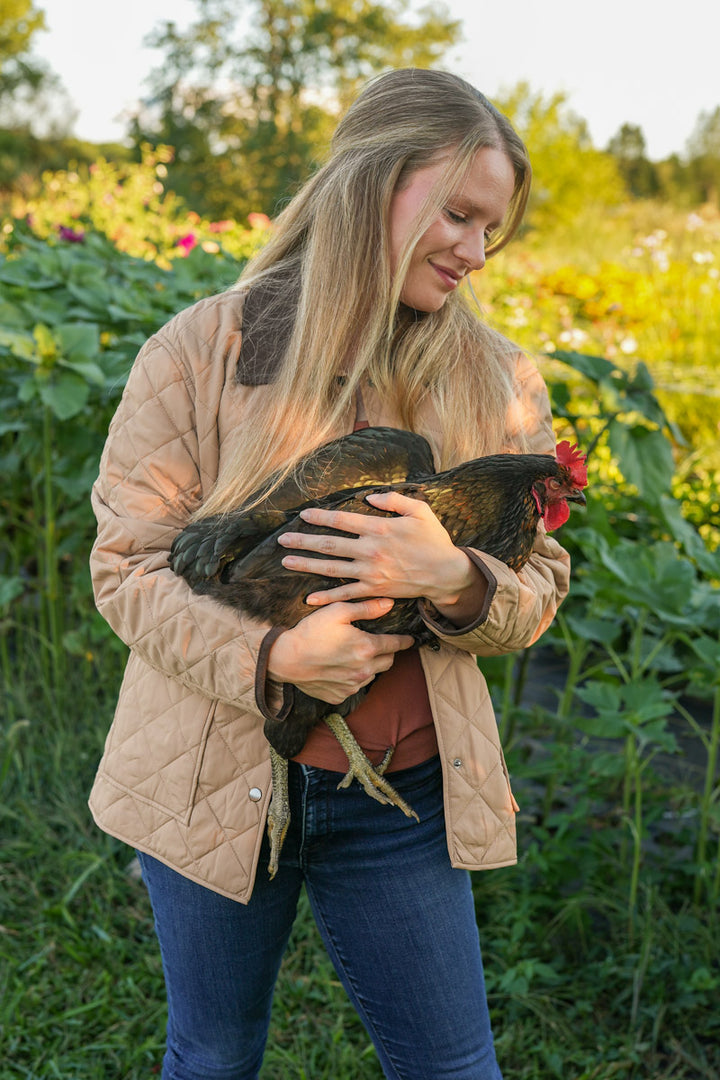 Woman holding a chicken in a field with greenery and flowers in the background