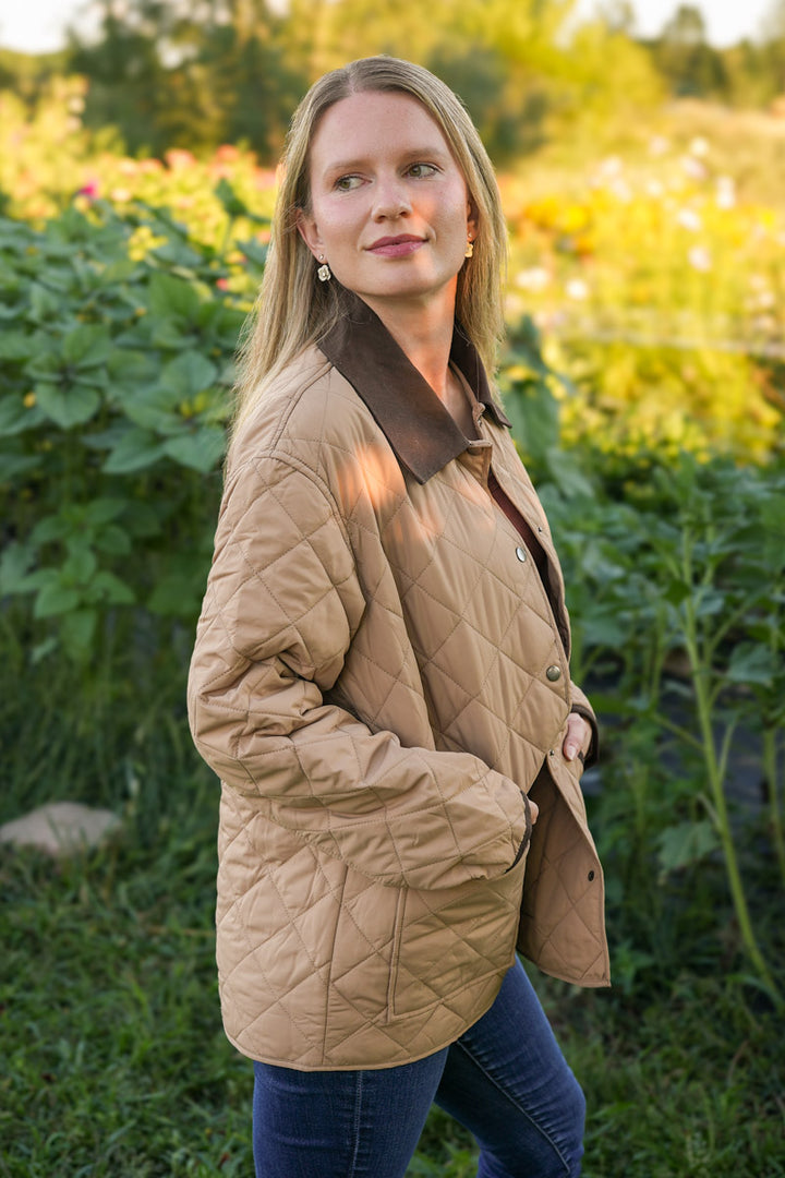 Woman wearing a beige quilted jacket with contrast collar standing in a field with greenery and flowers.