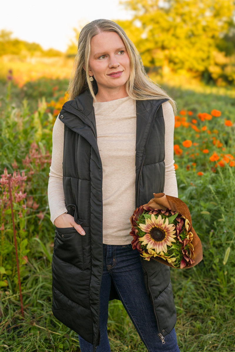 Woman wearing a long black puffer vest holding a bouquet of flowers in a field
