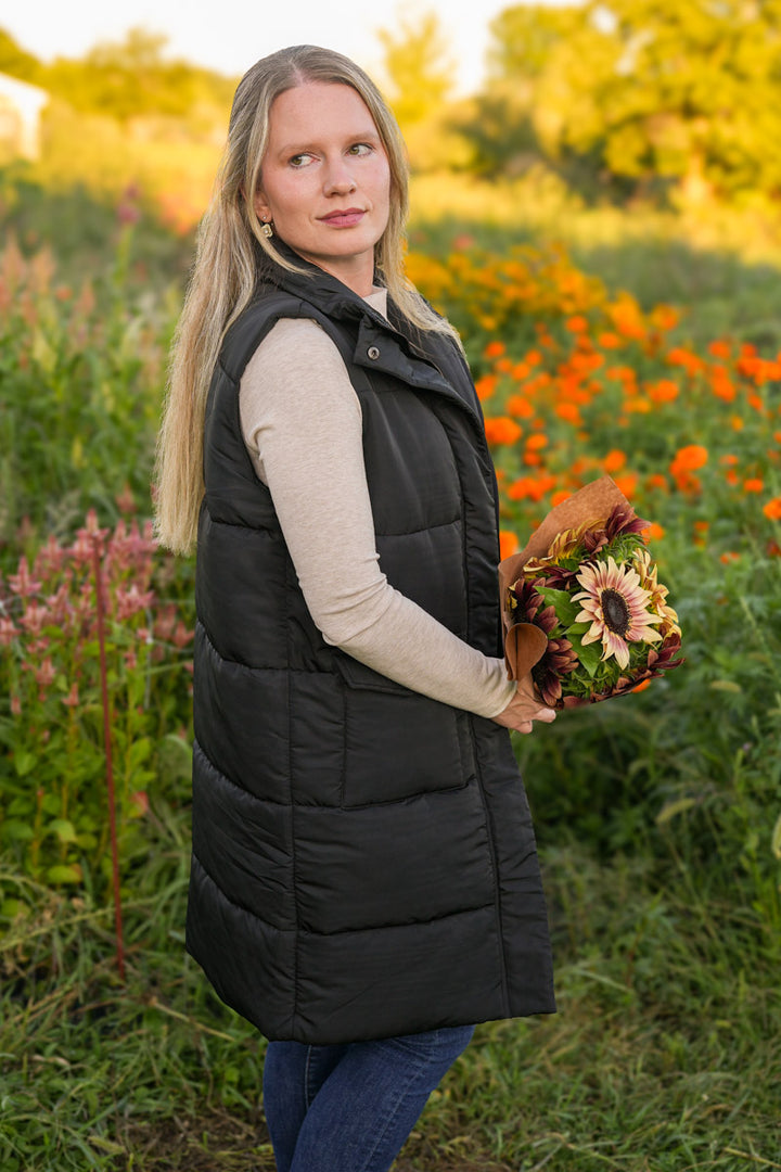 Woman wearing a long black puffer vest holding a bouquet of flowers in a field with a blurred background