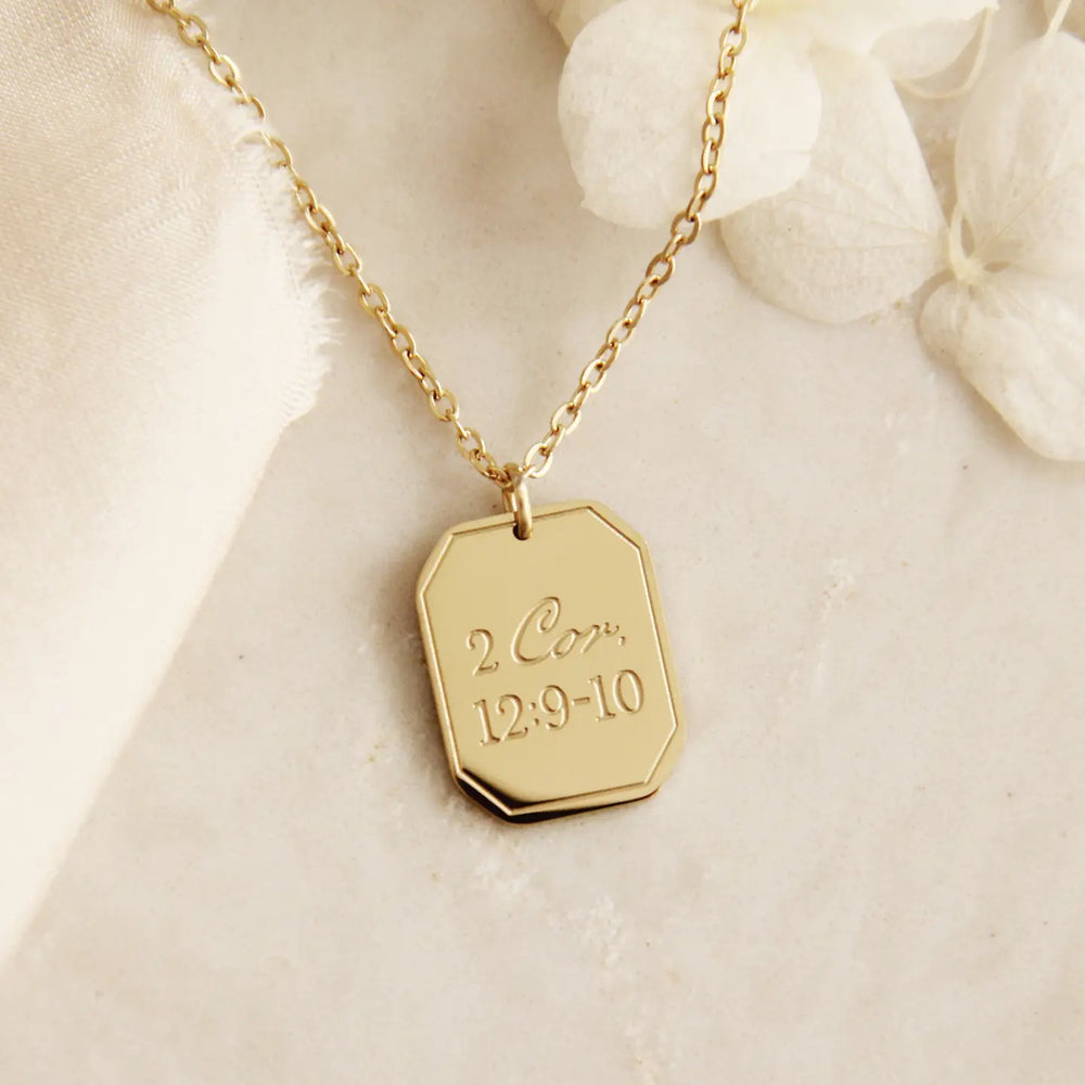 Gold Christian necklace with an engraved pendant that says 2 Cor. 12:9-10 on a soft beige background