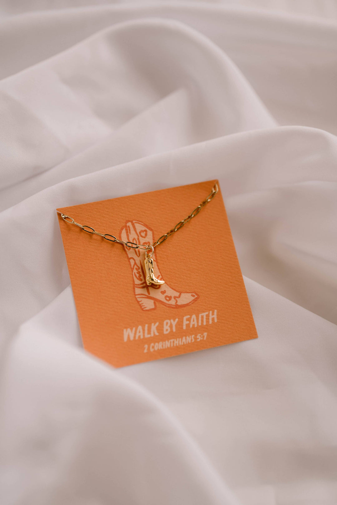 Gold necklace with a boot charm on an orange card with 'Walk by Faith' text, placed on a white fabric background.