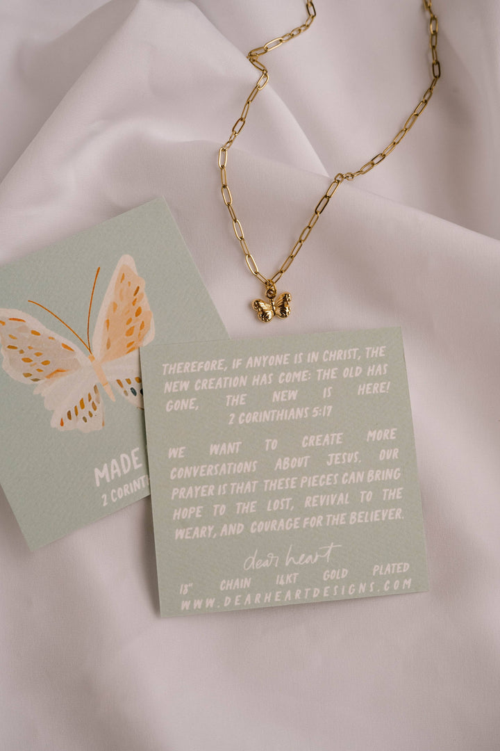 Gold butterfly necklace on a green card with text on a light pink fabric background