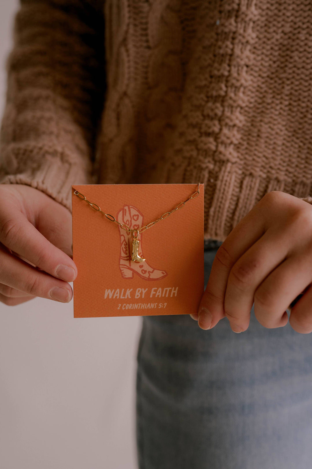 Person holding a necklace with a card that says 'Walk by Faith' against a neutral background