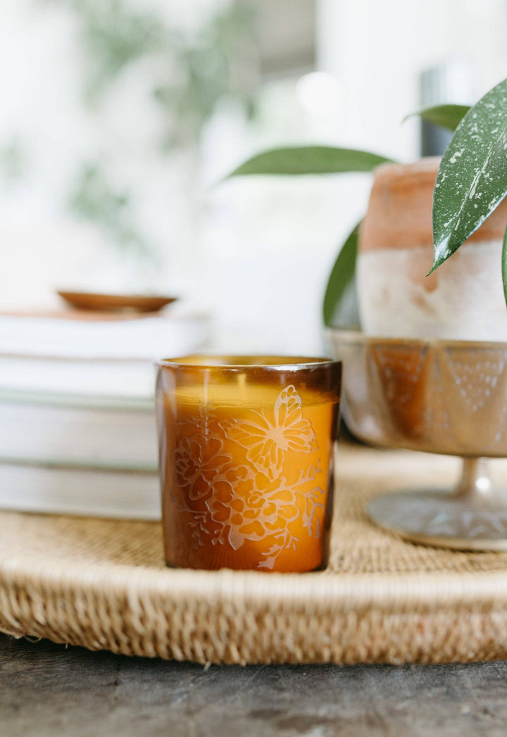 Decorative amber glass candle with floral design on a woven surface.