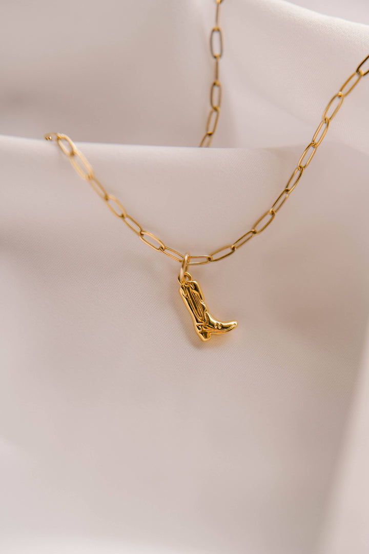 Gold necklace with a cowboy boot pendant on a light pink background