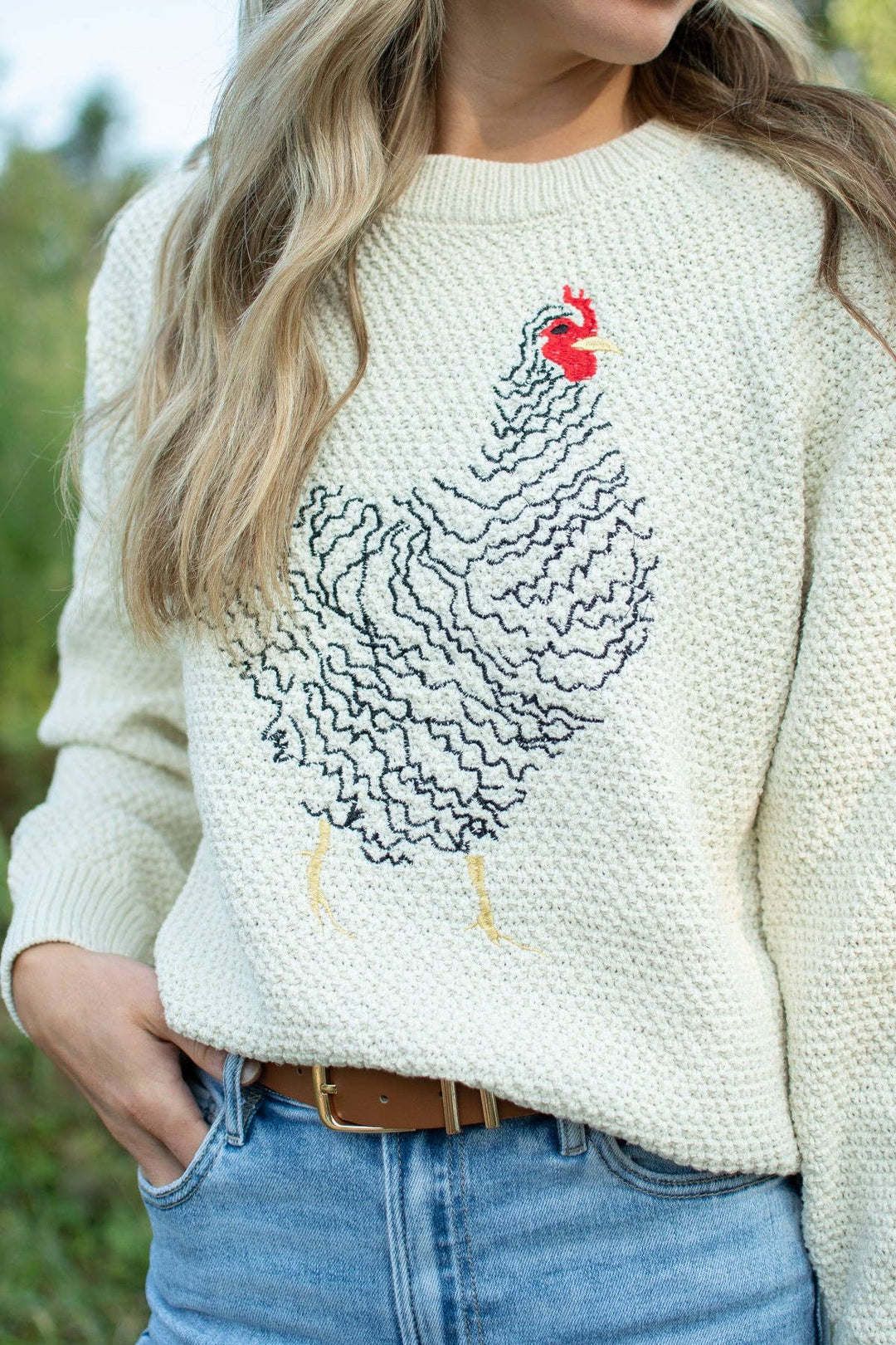 A woman wearing a cream-colored knit sweater with a large black wavy chicken design and a small red comb accent on the chest, paired with blue jeans.