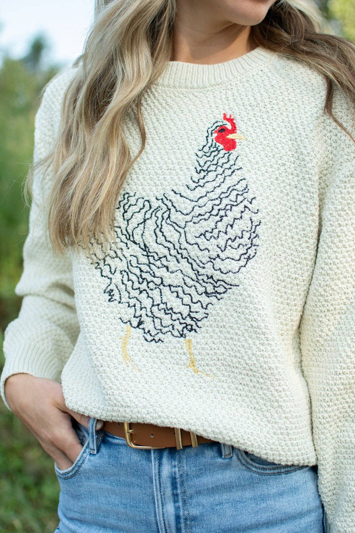 A woman wearing a cream-colored knit sweater with a large black wavy chicken design and a small red comb accent on the chest, paired with blue jeans.