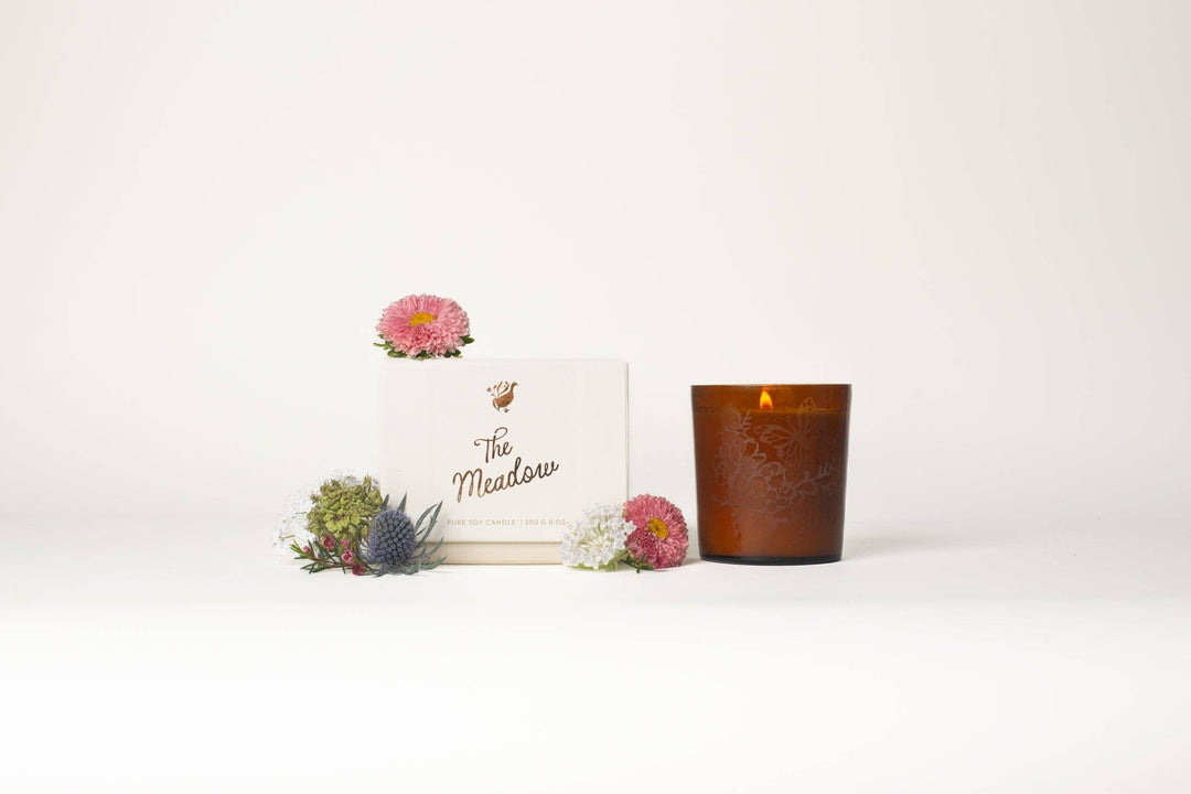 Candle and decorative box with 'The Meadows' branding on a white background