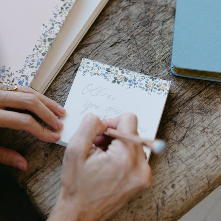Person writing on a small notepad with floral design on a wooden surface