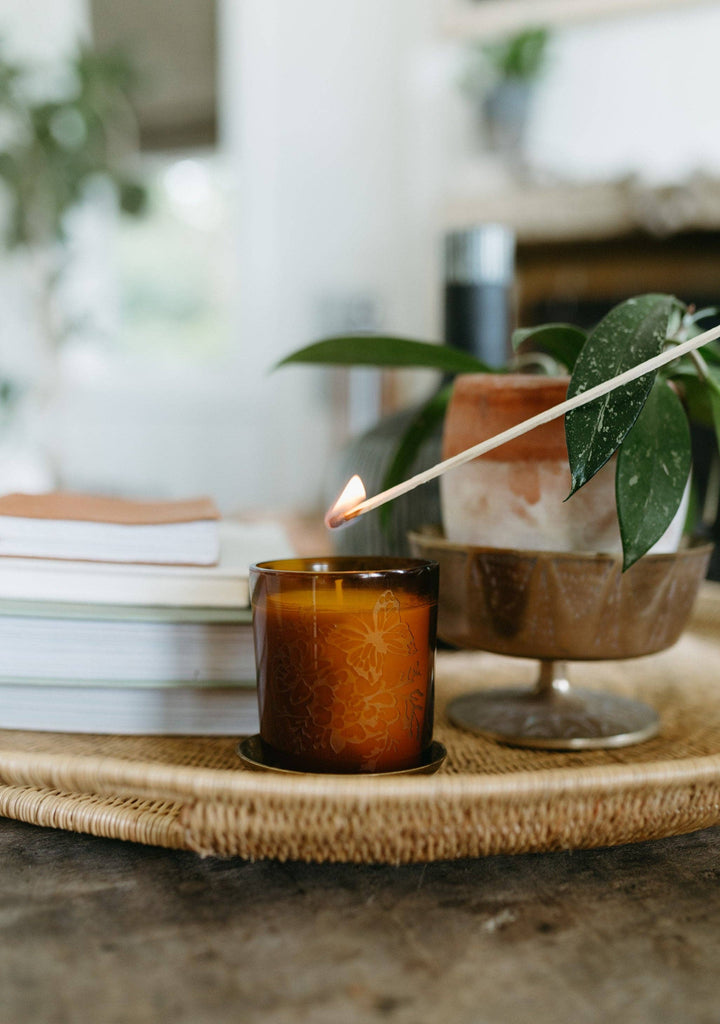 Candle being lit with a match on a woven tray with books and plants in the background