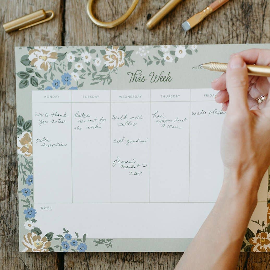 Weekly planner with floral design on a wooden surface, hand holding a pen.
