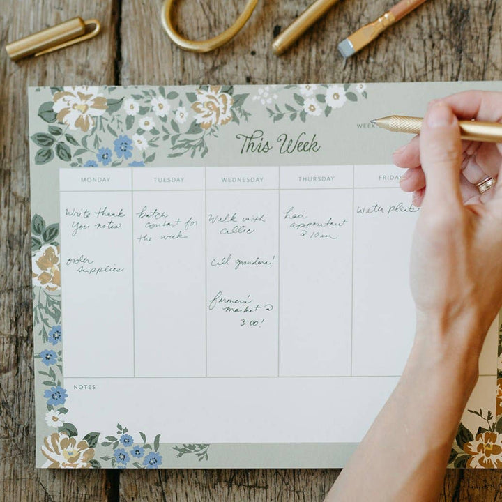 Weekly planner with floral design on a wooden surface, hand holding a pen.