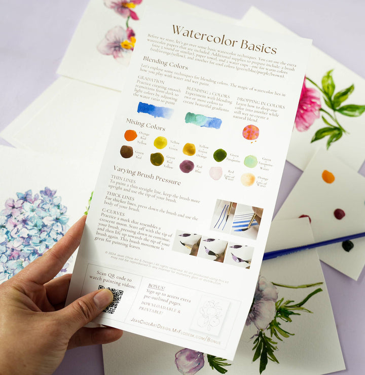 Hand holding a leaflet titled 'Watercolor Basics' with watercolor samples and instructions on a light purple background.