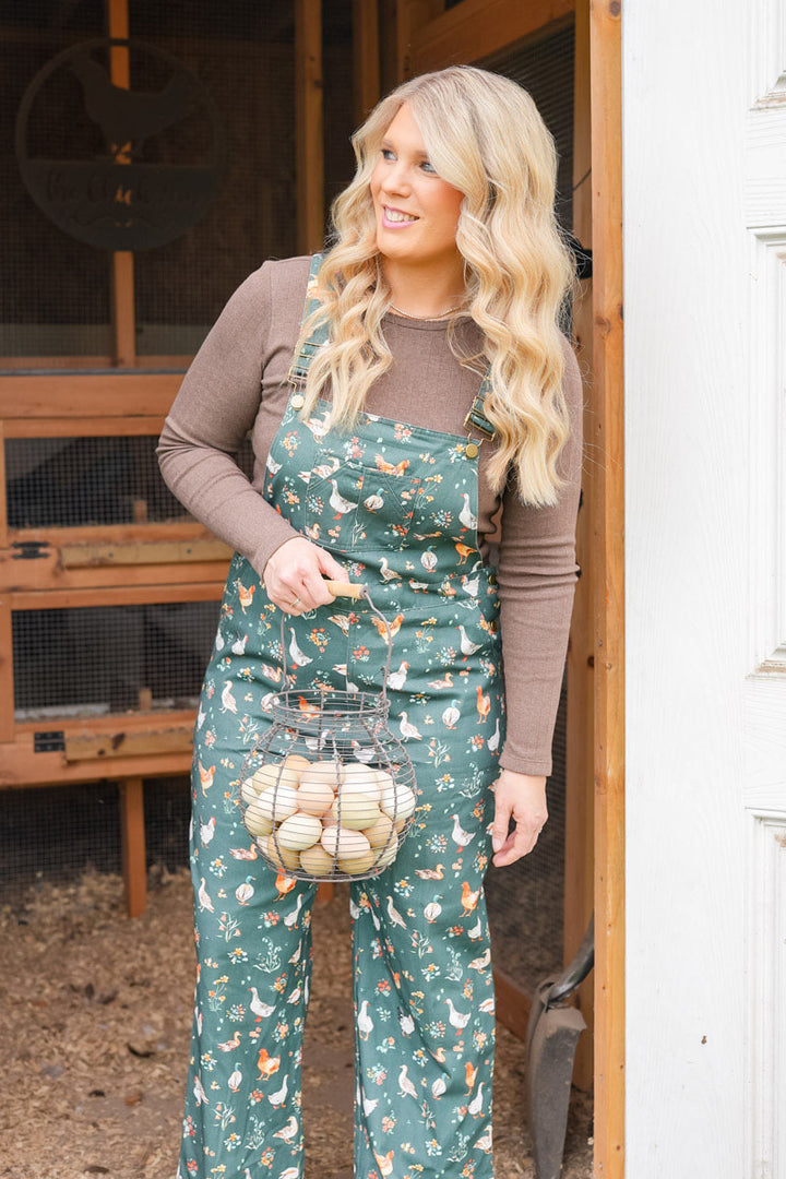A woman with long blonde hair stands in a doorway wearing forest green overall-style coveralls with a white duck and chicken print, layered over a brown long-sleeve shirt. She is holding a wire basket filled with eggs.
