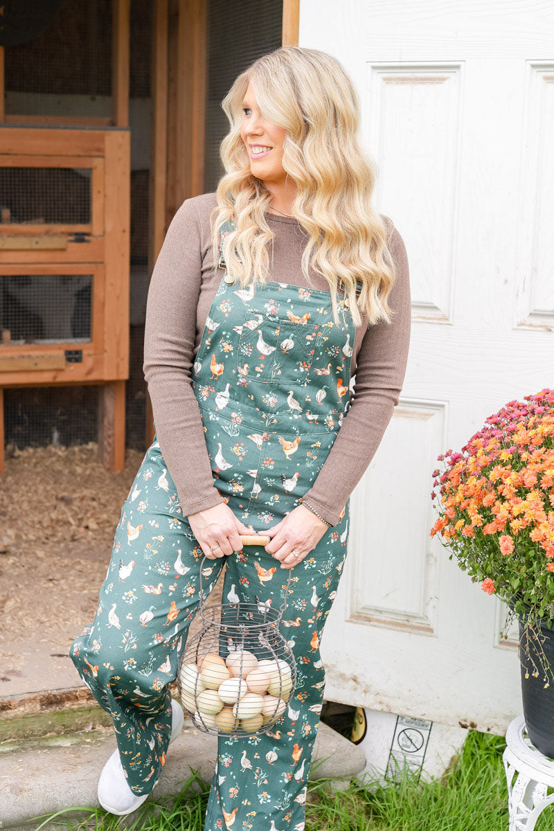 A woman stands outdoors in front of a white door and wooden coop, wearing green ducks and chickens patterned overalls over a brown long-sleeve shirt and holding a wire basket of eggs. A potted flower arrangement sits to the right.