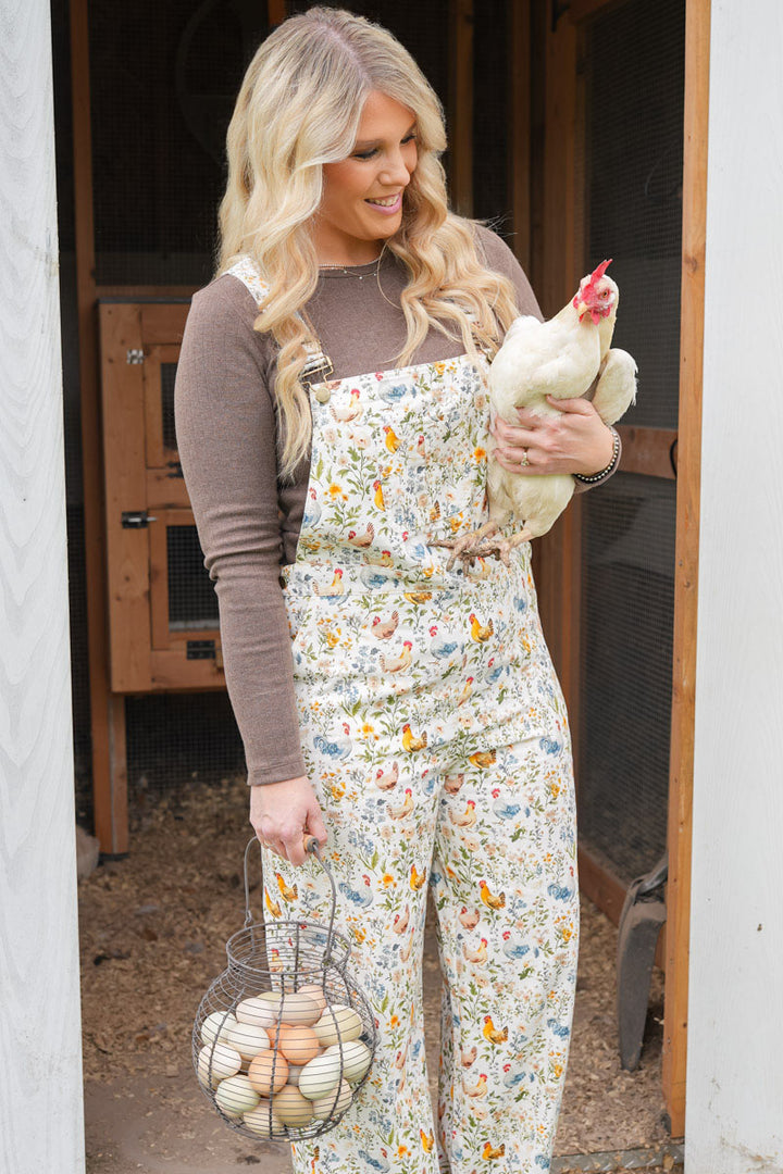 A woman stands in a doorway holding a white chicken and a wire basket of eggs, wearing floral print overalls over a brown long-sleeve shirt. The overalls feature a small multicolor pattern of chickens and flowers on a cream background.