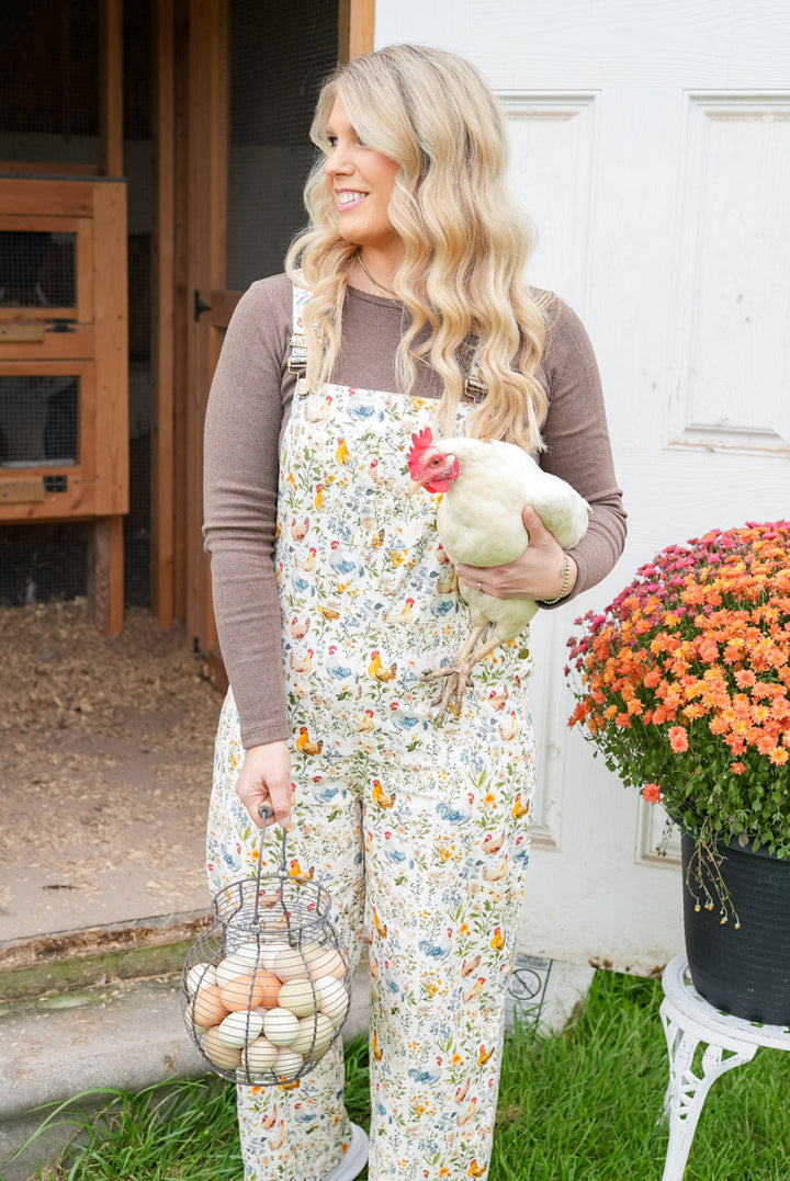 A woman stands on a farm porch wearing floral print overalls over a brown long-sleeve shirt, holding a live chicken and a wire basket of eggs. A white door, grass, and a potted flower are visible in the background.