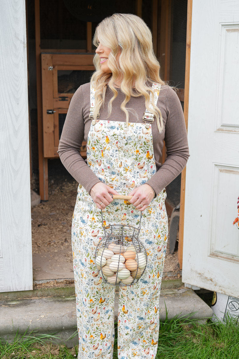 A woman stands in front of a white door and wooden barn doorway wearing cream floral overalls over a brown long-sleeve shirt, holding a wire basket filled with eggs.