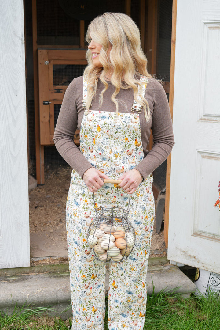 A woman stands in front of a white door and wooden barn doorway wearing cream floral overalls over a brown long-sleeve shirt, holding a wire basket filled with eggs.