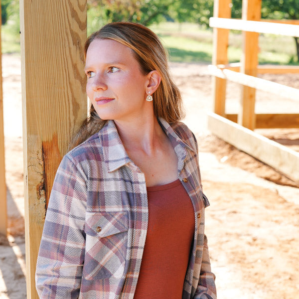 Woman wearing a plaid shirt and brown top standing outdoors near wooden structures.