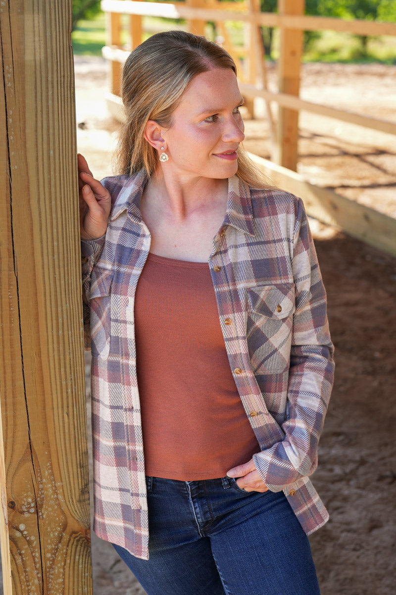 Woman wearing a plaid shirt over a brown top and jeans, standing against a wooden fence.