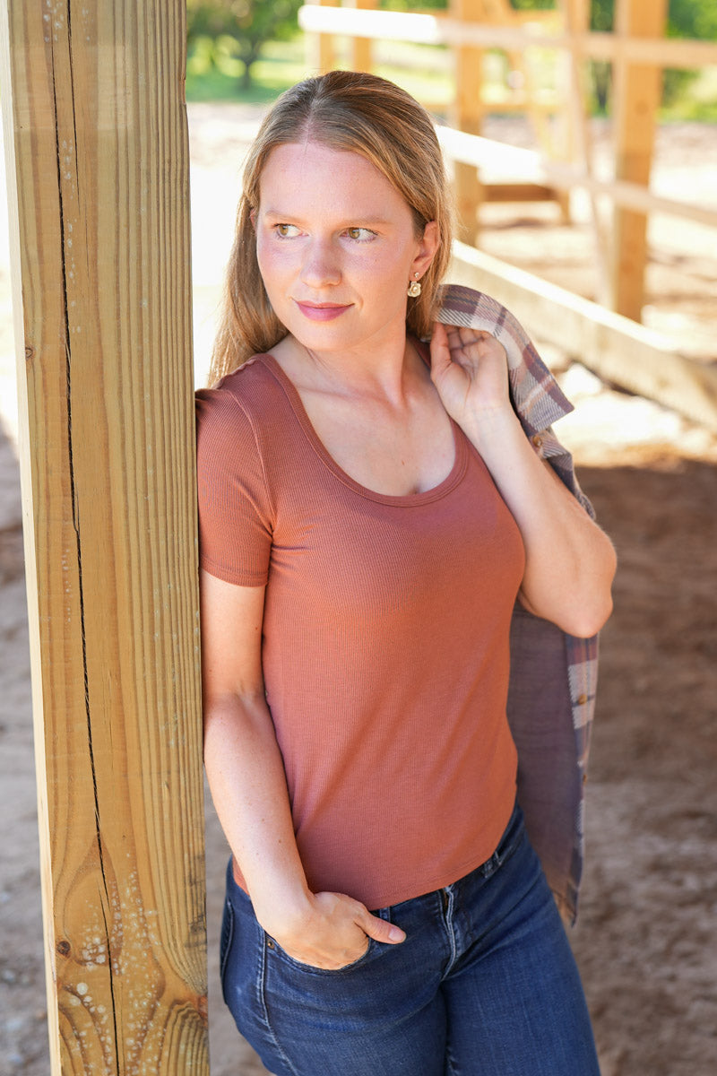 Woman in a barn wearing a rust-colored shirt and blue jeans, with a plaid shirt over her shoulder.