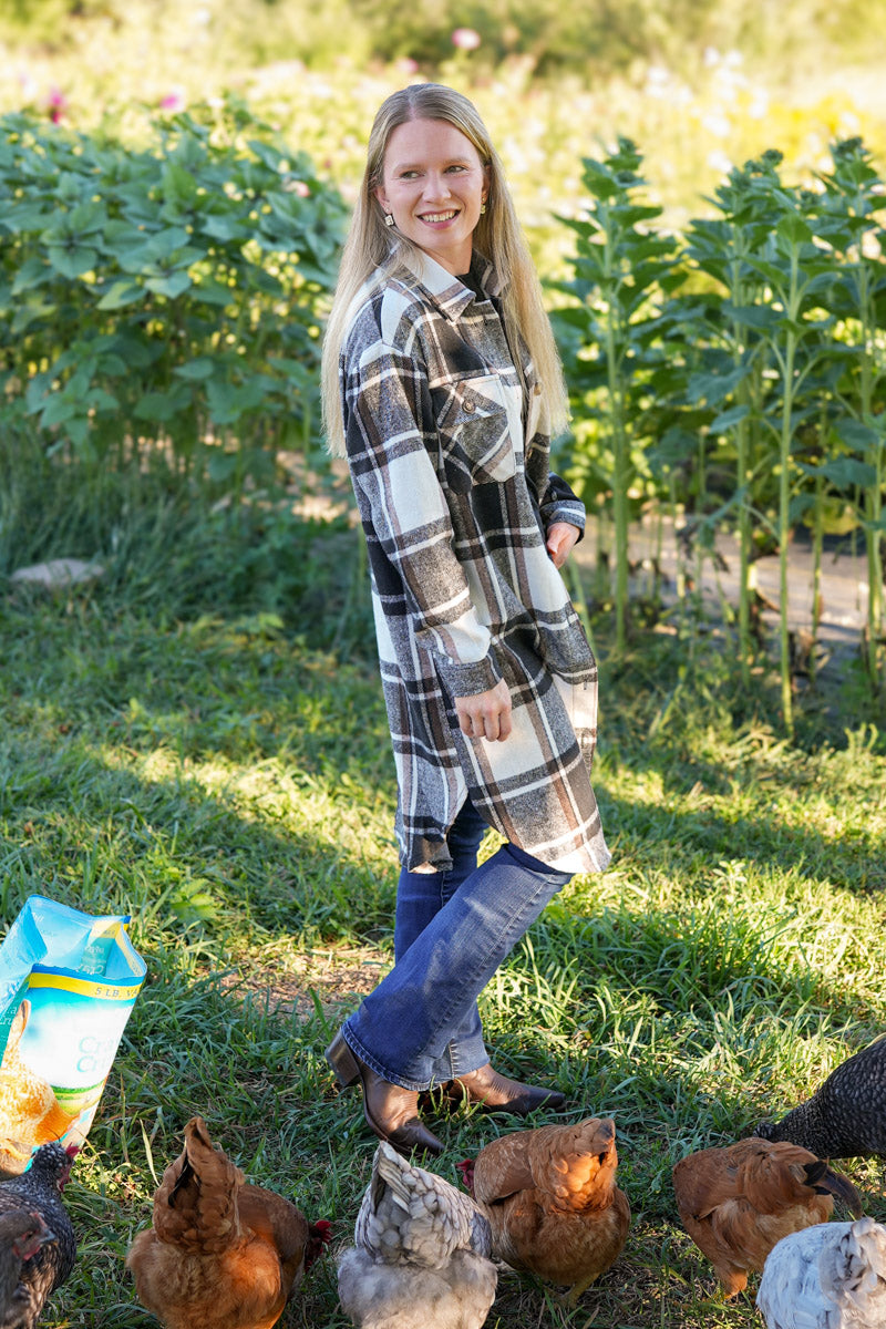 Woman in a long plaid shirt and jeans standing in a garden with chickens around her