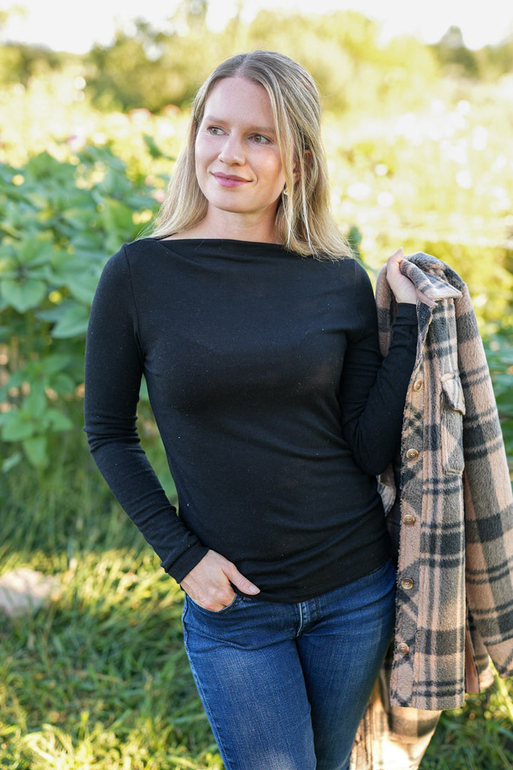 Woman wearing a black long-sleeve top and blue jeans, holding a plaid jacket outdoors.