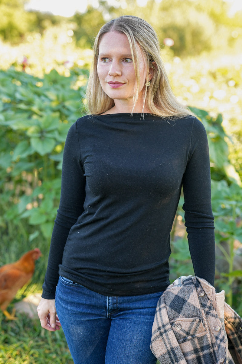Woman wearing a black long-sleeve top and blue jeans standing outdoors with greenery and a chicken in the background.