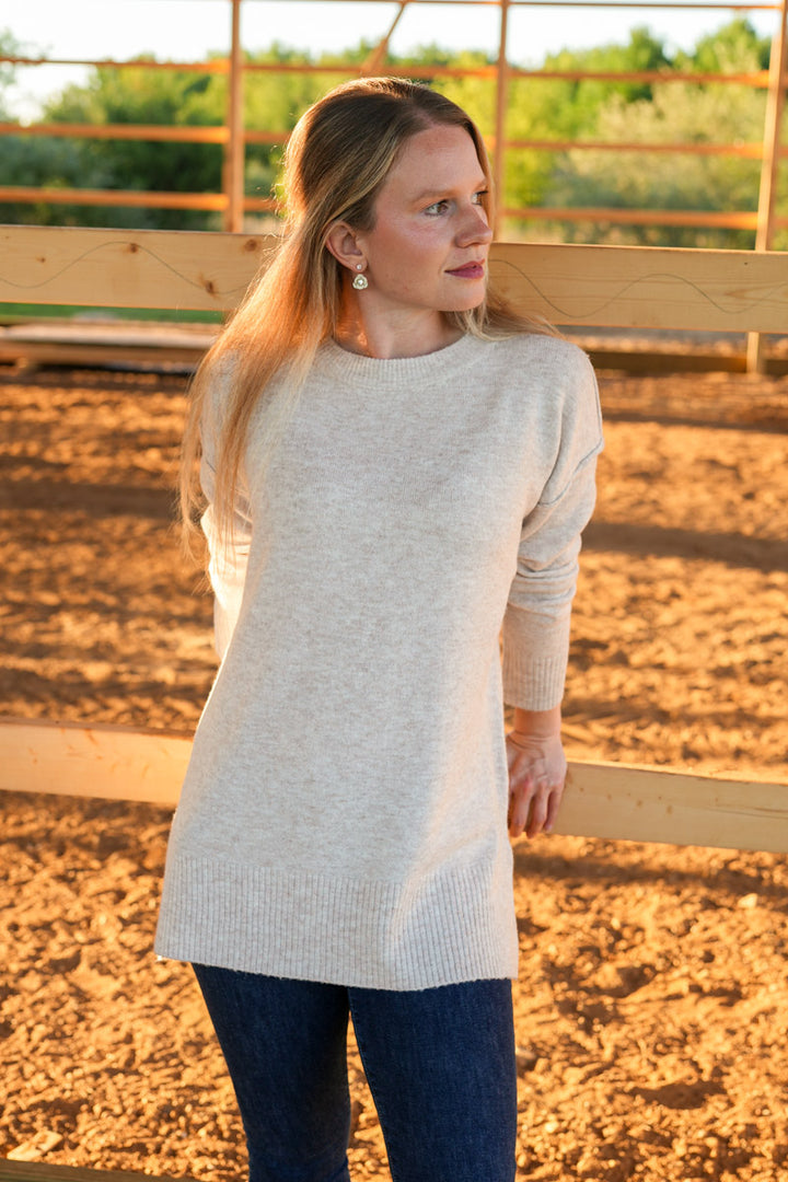 Woman wearing a light taupe sweater and dark blue jeans standing in an outdoor setting with wooden fencing and greenery.