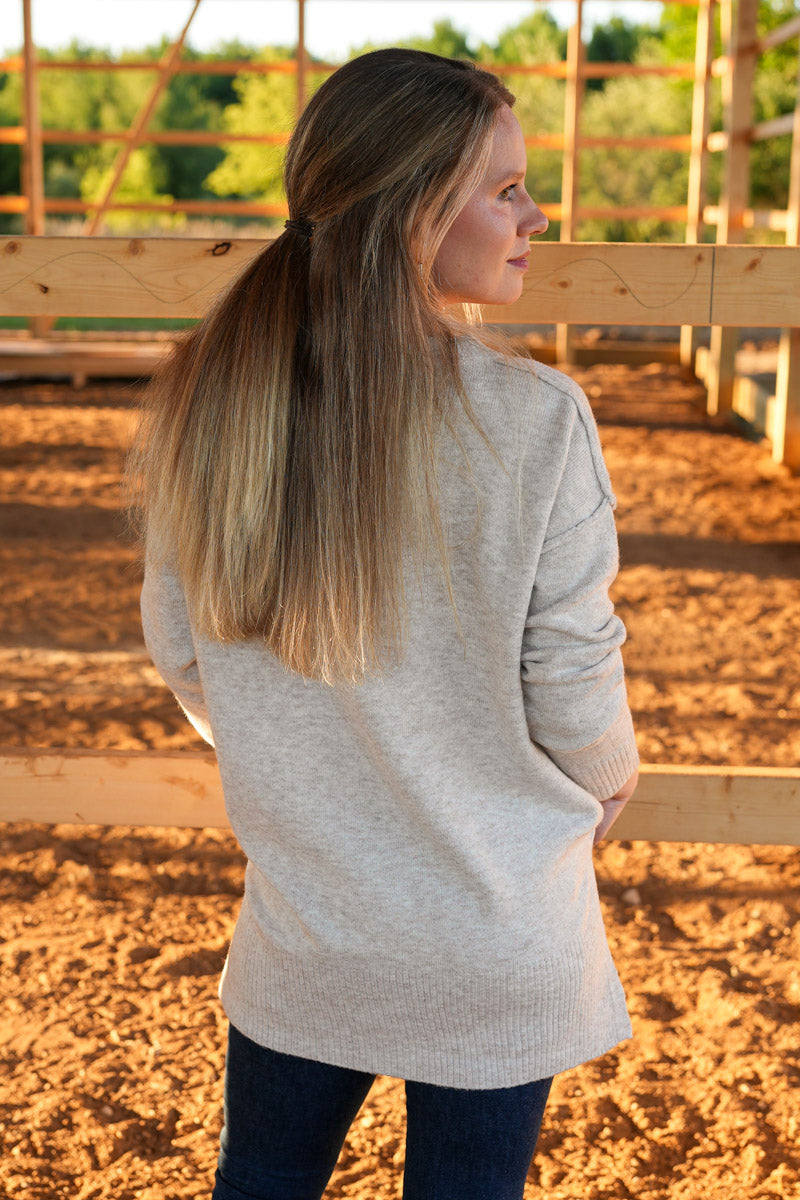 Woman wearing a light taupe sweater standing in an outdoor setting with wooden fences and trees.
