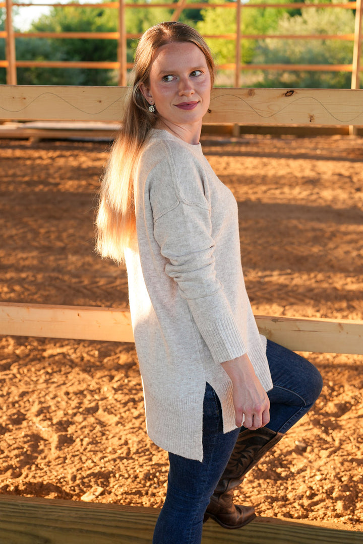 Woman wearing a long, taupe crewneck sweater standing by a wooden frame