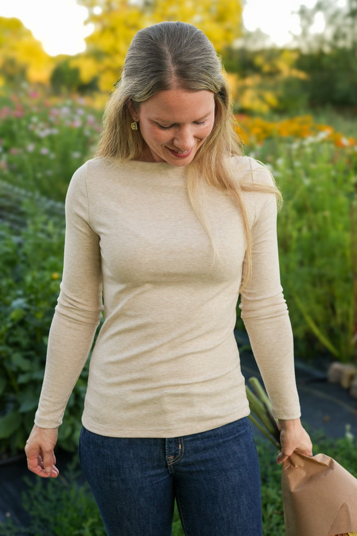 Woman in a beige long-sleeve top and blue jeans standing in a garden.