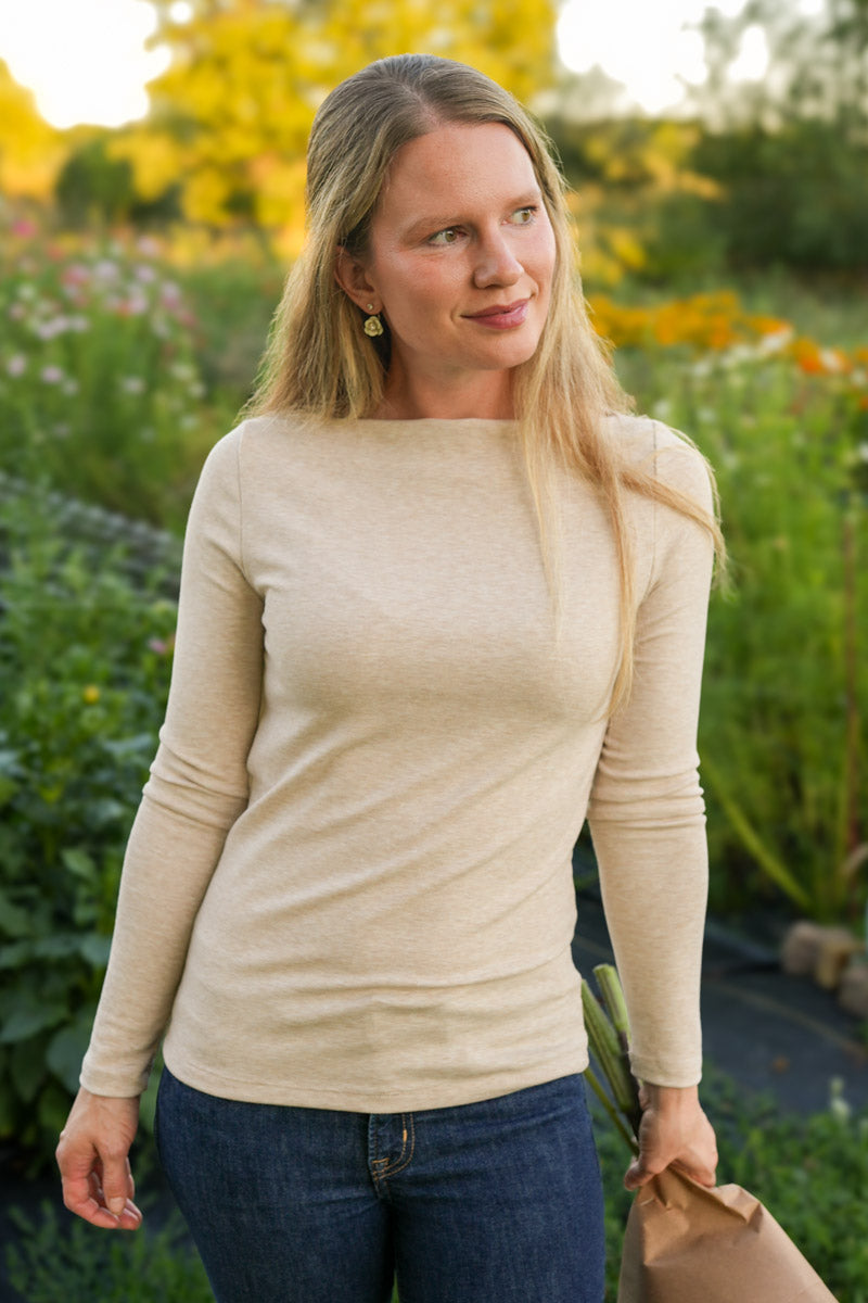 Woman wearing a taupe long sleeve boat neck top standing in a garden with flowers and greenery.