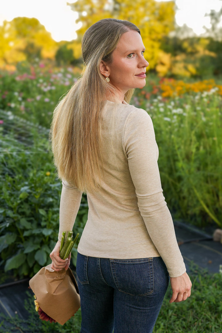 Woman standing in a garden holding flowers, wearing a long sleeve taupe top and blue jeans.