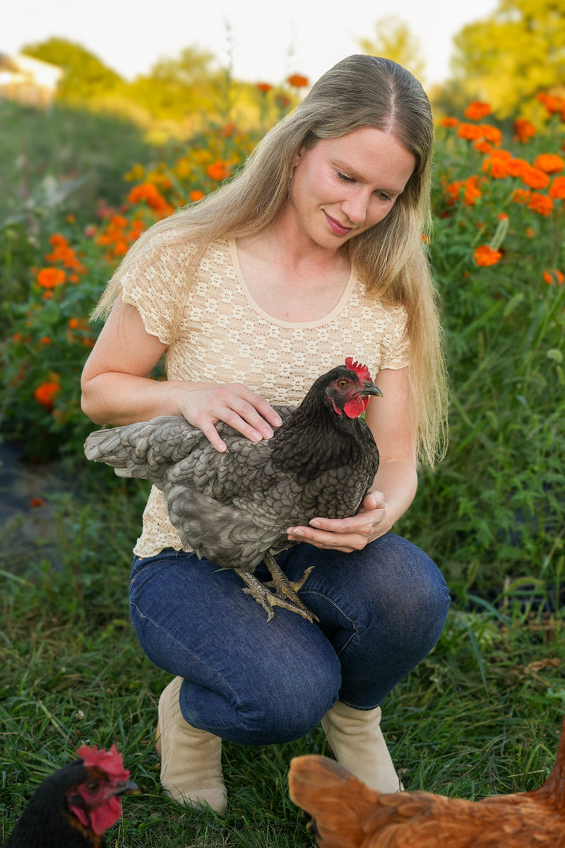 Woman holding a chicken in a garden with flowers and other chickens in the background