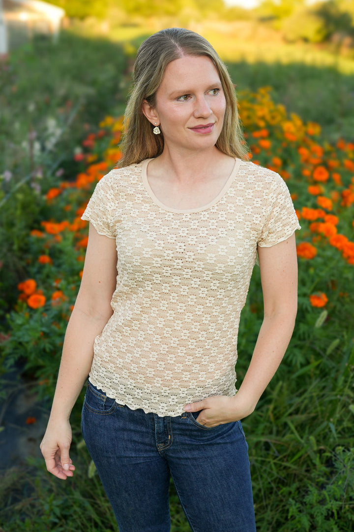 Woman wearing a floral lace beige top and blue jeans standing in a garden with orange flowers.