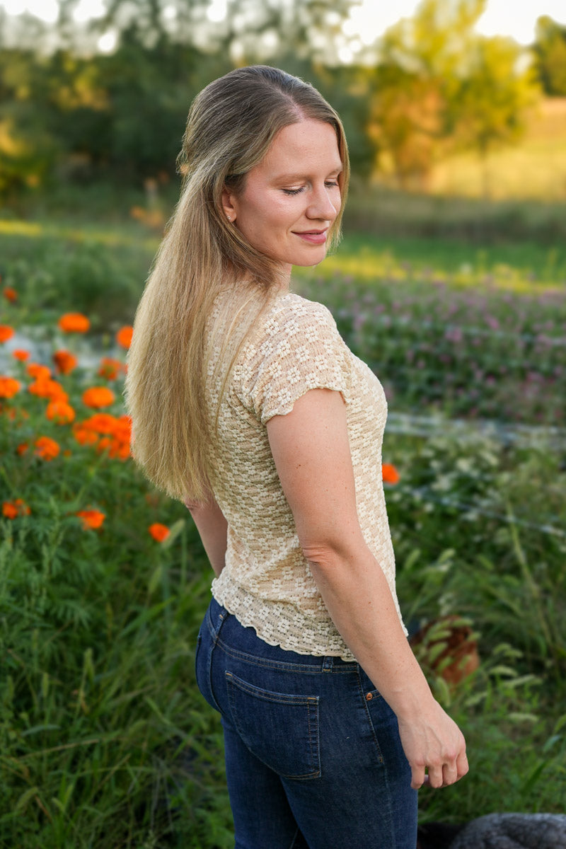 Woman standing in a field of flowers wearing a beige top and blue jeans.