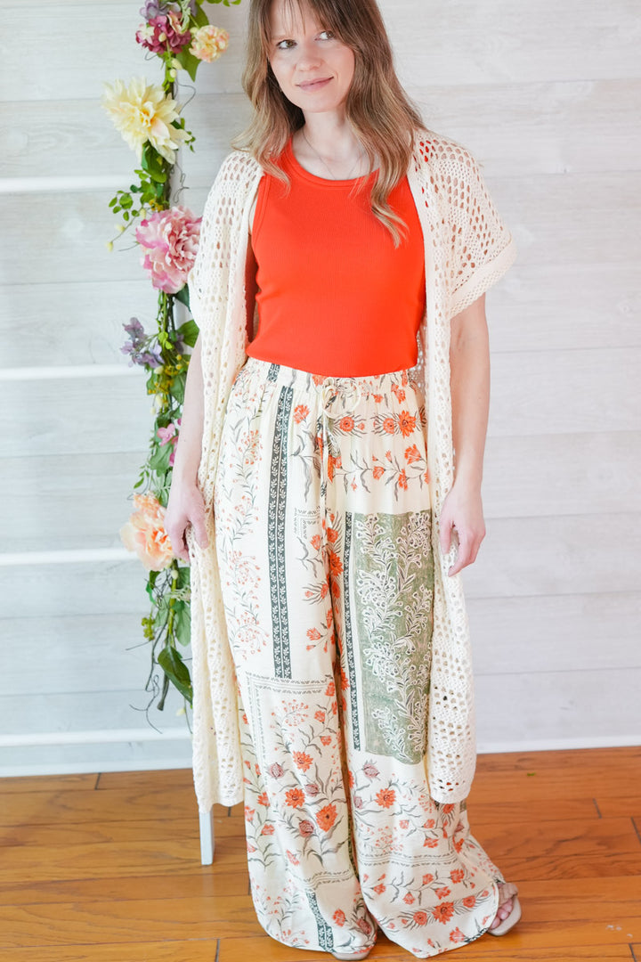 Woman wearing a red top and vintage floral pants with a white lace cardigan, standing against a white wall with flowers.