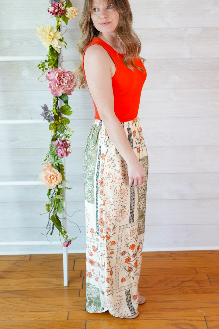 Woman wearing a red tank top and vintage floral patterned pants standing next to a floral arrangement indoors.