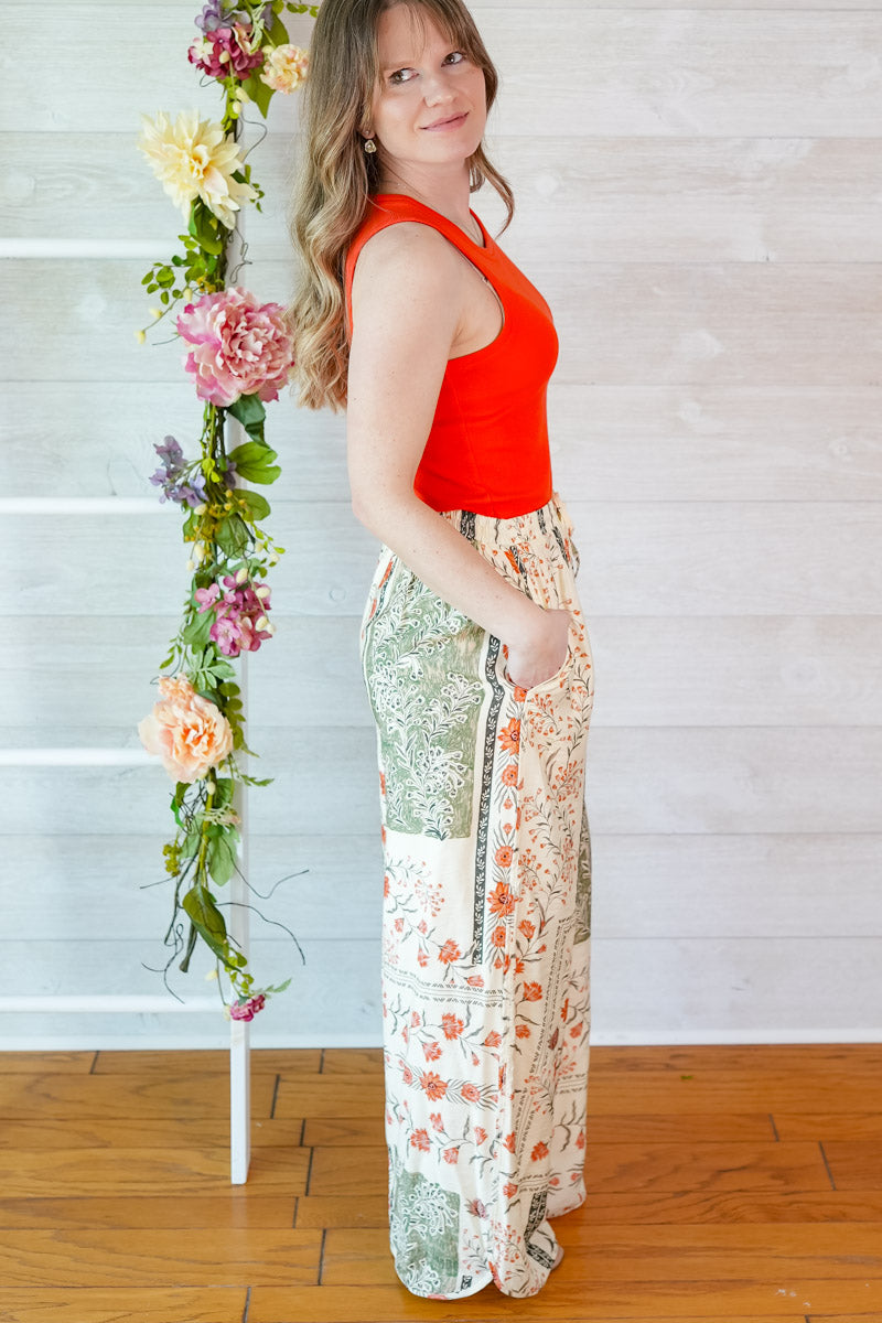 Woman in a red tank top and vintage floral patterned pants standing next to a floral arrangement indoors.