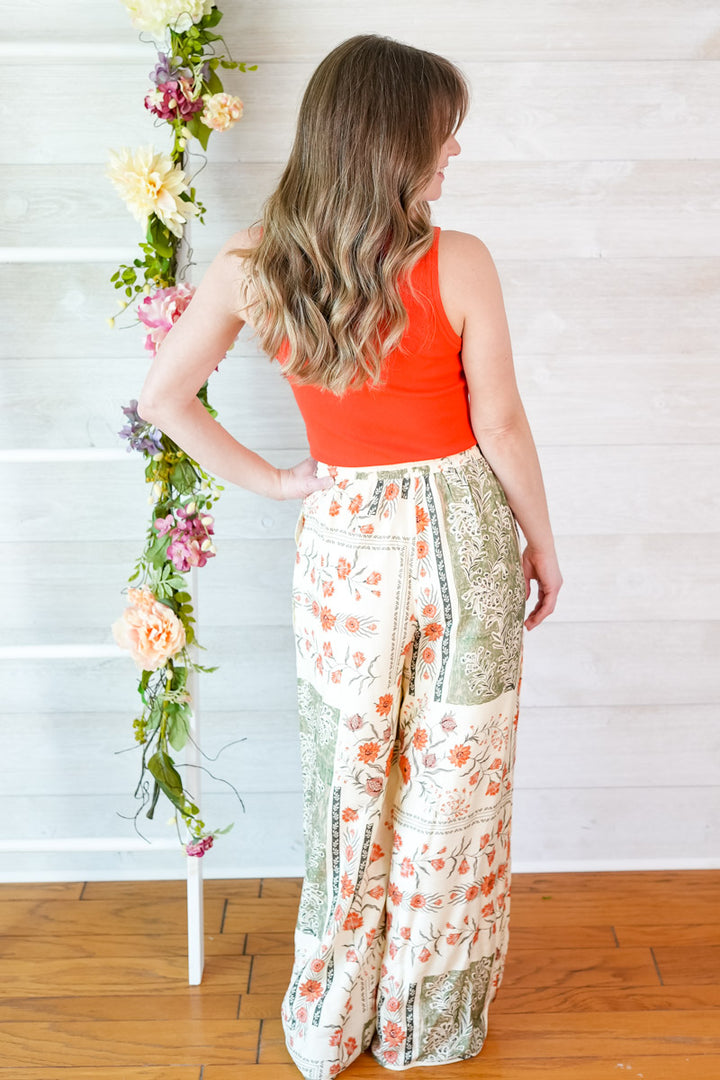 Woman wearing a bright red orange tank top and vintage botanical floral pants standing next to a floral arrangement against a white wall.